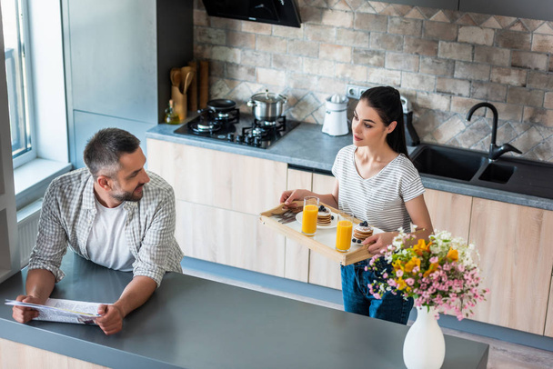 Mann mit Zeitung steht am Tresen und schaut Frau mit Frühstück auf Holztablett in Küche an - Foto, Bild