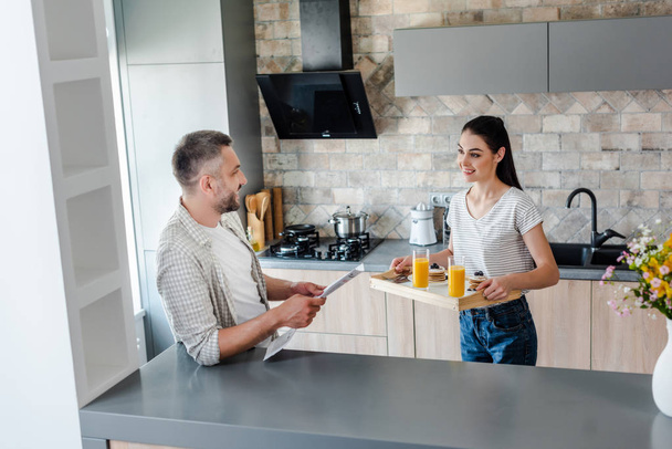 muž s novinami stojí u přepážky a při pohledu na ženu se snídaní na dřevěný tác v kuchyni - Fotografie, Obrázek