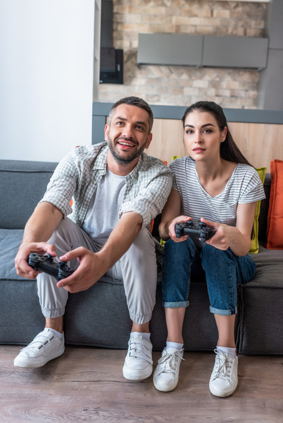 married couple with gamepads playing video games together while sitting on sofa at home - Fotó, kép