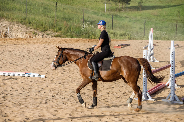 Foto al aire libre de la mujer rubia belleza montar a caballo durante el día. Mujer joven en ropa deportiva disfrutando de la equitación en el hipódromo
  - Foto, imagen
