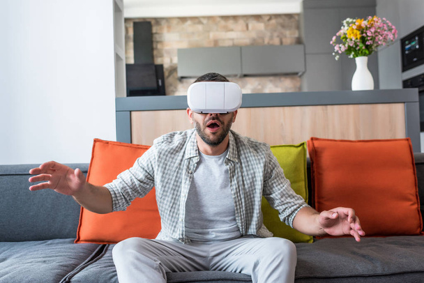 emotional man wearing virtual reality headset while sitting on sofa at home - Foto, afbeelding