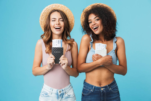Image of two happy vacation girls 20s with different color of skin in straw hats and swimsuits smiling while holding passports with travel tickets isolated over blue background - Foto, Imagem