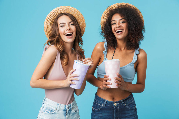 Photo of two joyful multiethnic women 20s wearing straw hats and summer clothing laughing while drinking soda from big paper cups isolated over blue background - Photo, image