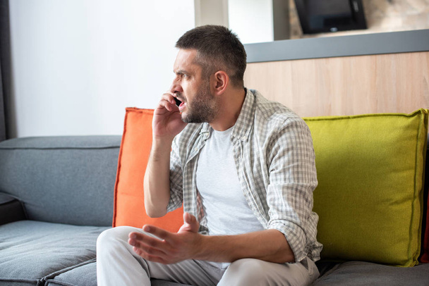 emotional bearded man sitting on sofa and talking on smartphone at home - Photo, Image