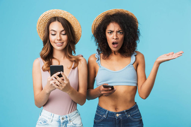 Photo of angry african american woman holding mobile phone and expressing outrage, while her female friend using smartphone with pleasure isolated over blue background - Foto, immagini