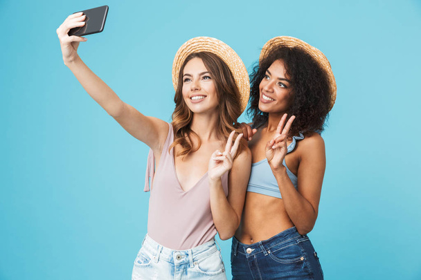 Two multiethnic holiday women wearing straw hats smiling and showing peace sign at camera, while taking selfie photo on smartphone isolated over blue background - Zdjęcie, obraz