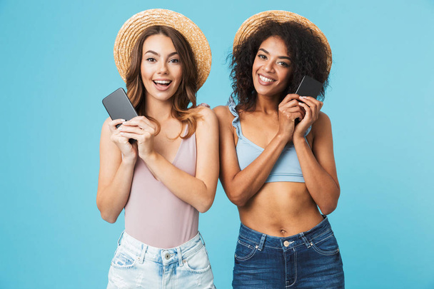 Dwie piękne kobiety wieloetnicznym 20s sobie słomkowe kapelusze, uśmiechając się i obie smartphone gospodarstwa patrząc na kamery na białym tle niebieski - Zdjęcie, obraz