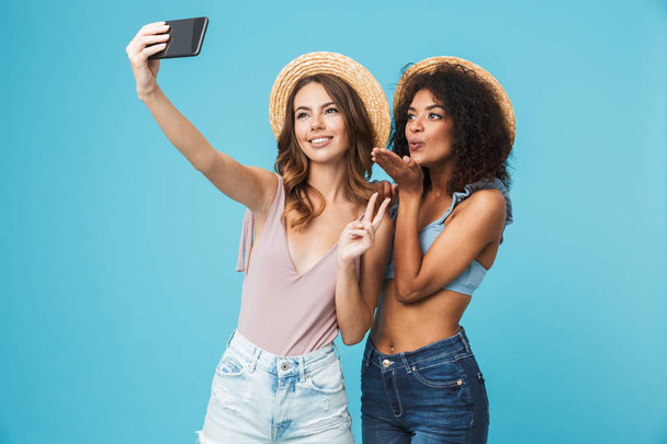 Vacation photo of two multiethnic girls wearing straw hats smiling and showing peace sign at camera, while taking selfie on smartphone isolated over blue background - 写真・画像
