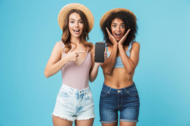 Amusing photo of caucasian and african american women expressing positive emotions while pointing finger at copyspace screen of cell phone isolated over blue background - 写真・画像