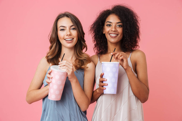 Image of cheerful young two women standing isolated over pink background drinking cocktails. - Foto, Imagem