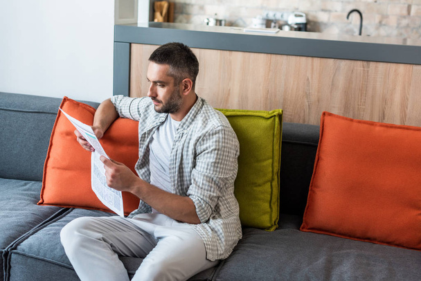 portrait of focused man reading newspaper on sofa at home - Фото, изображение