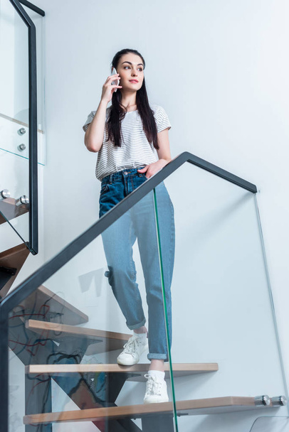 low angle view of attractive woman talking on smartphone while standing on stairs at home - Foto, Imagem