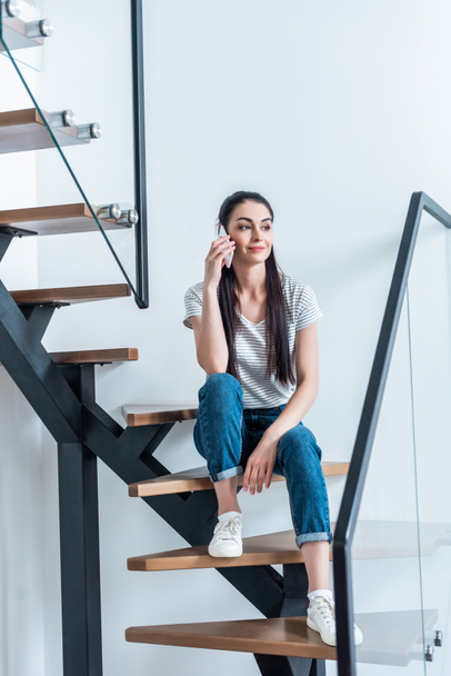 smiling woman sitting on stairs and talking on smartphone at home - Foto, Imagem
