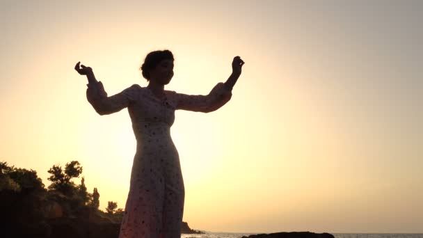 Очаровательная кавказская брюнетка в светлом платье танцует на скале над морем на восходе солнца
 - Кадры, видео