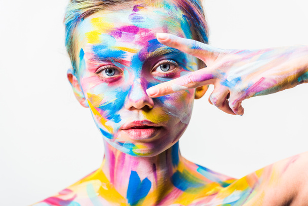 ελκυστική κοπέλα με πολύχρωμο φωτεινό σώμα τέχνη βλέπουν κάμερα μέσα από δύο δάχτυλα που απομονώνονται σε λευκό   - Φωτογραφία, εικόνα