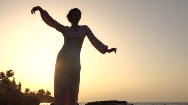 Rozkošný kavkazské bruneta dívka ve světle šaty tančí na útesu nad mořem v době východu slunce - Záběry, video