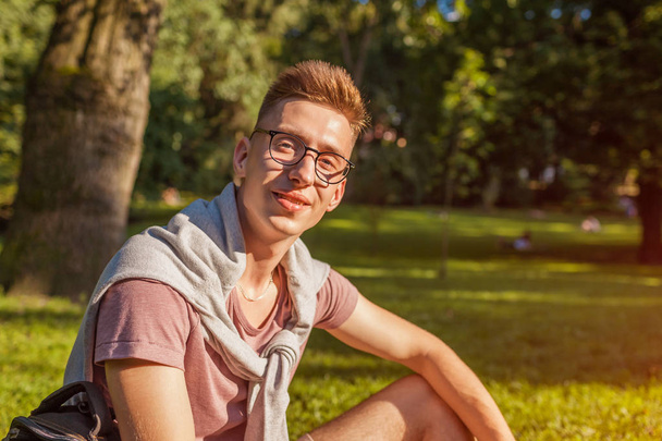 stattliche College-Mann beim Chillen im Frühling Campus Park. Glücklicher Kerl Student sitzt auf Gras und schaut in die Kamera bei Sonnenuntergang - Foto, Bild