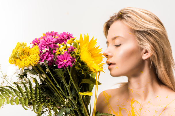 side view of attractive woman with yellow paint on body sniffing flowers isolated on white - 写真・画像