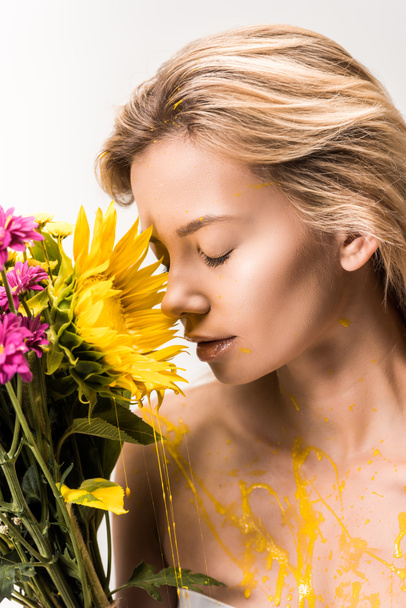 attractive woman with yellow paint on body sniffing bouquet of flowers isolated on white - Фото, зображення