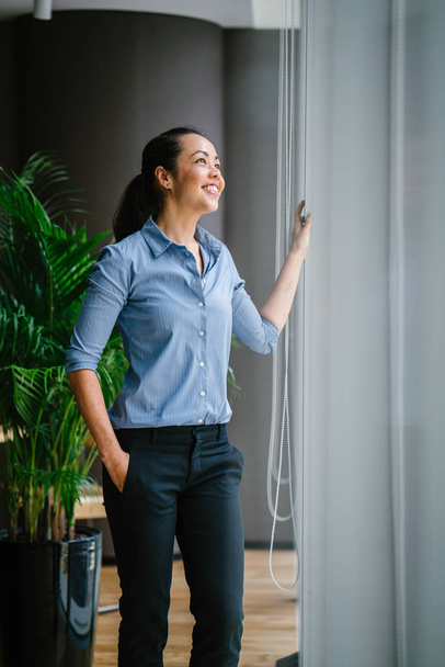 Portrait of young confident asian businesswoman wearing blue shirt smiling standing near window - Фото, изображение
