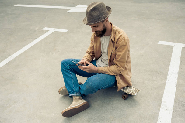 wysoki kąt widzenia stylowe Brodaty mężczyzna siedzi na deskorolce i przy użyciu smartfona  - Zdjęcie, obraz