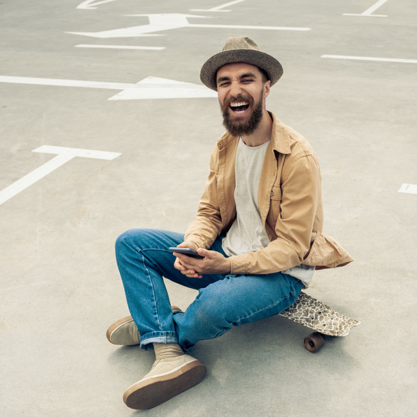 happy bearded man sitting on skateboard and using smartphone - Φωτογραφία, εικόνα
