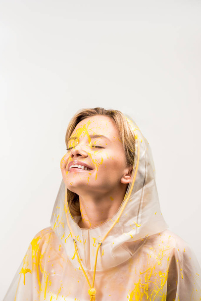 happy beautiful woman in raincoat painted with yellow paint standing with closed eyes isolated on white  - Photo, Image
