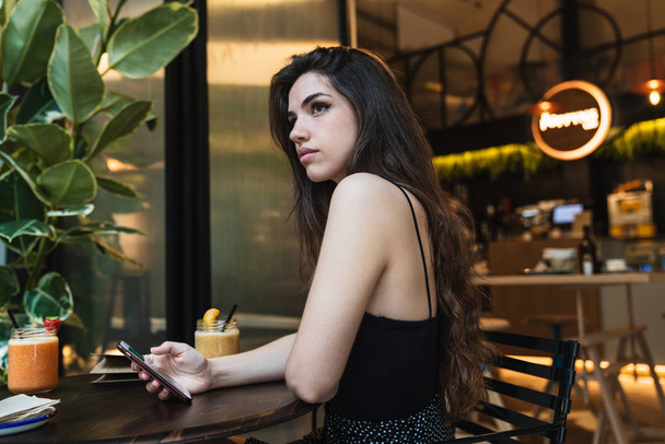 Side view of charming young lady holding smartphone and looking away while sitting at table in stylish cafe - Foto, Imagen