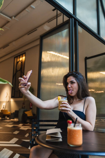 Attractive young female holding glass of yummy smoothie and showing tongue while taking selfie at table in stylish cafe - Zdjęcie, obraz