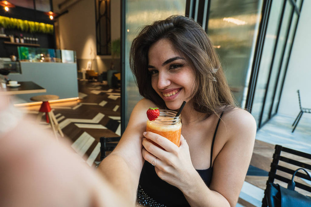 Menina jovem atraente com vidro de bom smoothie sorrindo e olhando para a câmera enquanto toma selfie no café elegante
 - Foto, Imagem