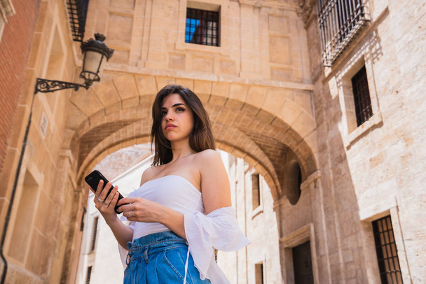 Pretty young lady in casual outfit holding smartphone while standing on beautiful street of Valencia in Spain - Фото, зображення