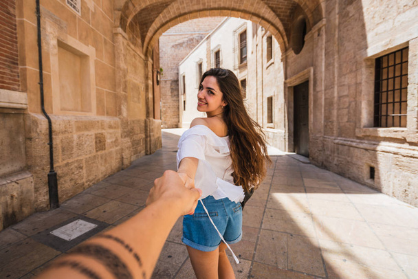 Beautiful young brunette smiling and leading anonymous friend by hand while walking on amazing street on sunny day in Valencia - Фото, зображення