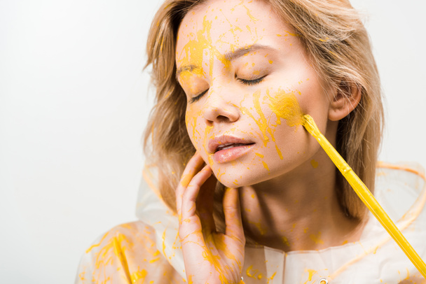 headshot of beautiful woman in raincoat painting face with yellow paint isolated on white  - Photo, Image