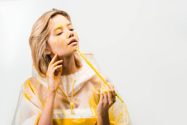 beautiful woman in raincoat painting face with yellow paint isolated on white  - Zdjęcie, obraz