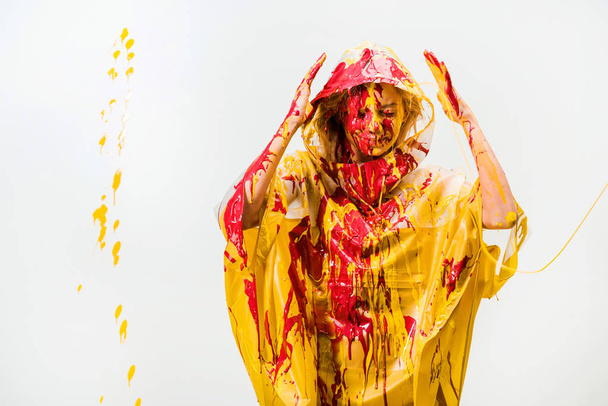beautiful woman in raincoat painted with yellow and red paints protecting head with hands isolated on white - Photo, Image