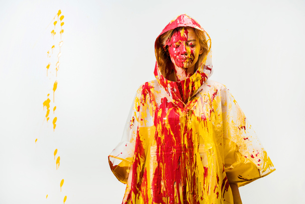 attractive woman in raincoat painted with yellow and red paints standing with closed eyes isolated on white  - Φωτογραφία, εικόνα