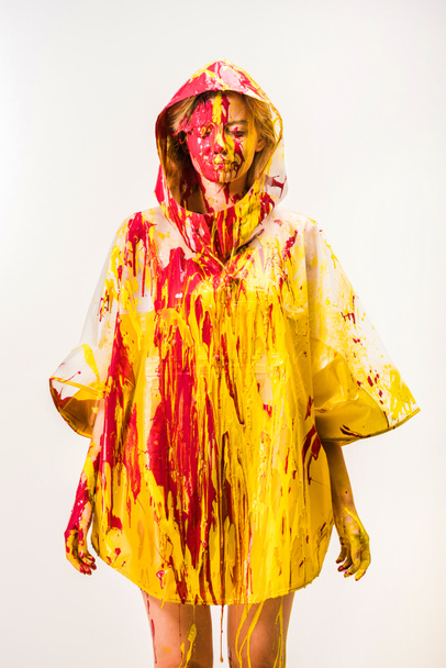 beautiful woman in raincoat painted with yellow and red paints standing with closed eyes isolated on white  - Fotó, kép