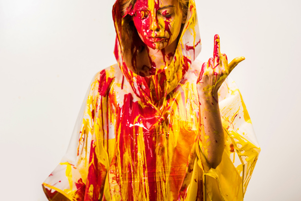 γυναίκα σε αδιάβροχο βαμμένες με κίτρινα και κόκκινα χρώματα που δείχνει το μεσαίο δάκτυλο απομονωθεί σε λευκό  - Φωτογραφία, εικόνα