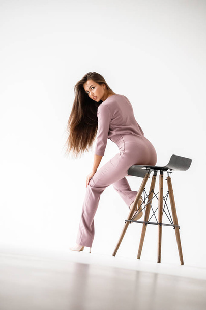Portrait of beautiful brunette woman wearing casual purple suit, sitting on wooden chair, white studio background - Photo, Image