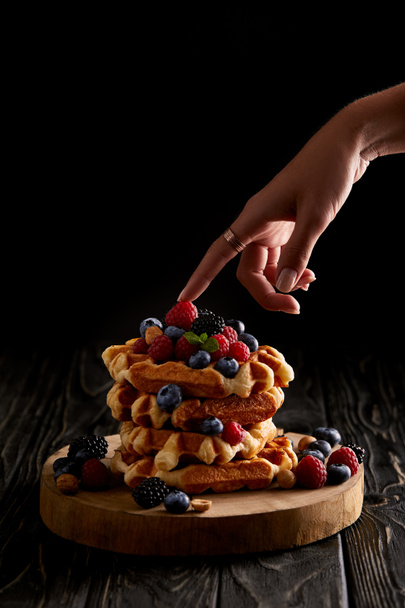 tiro cortado de mulher tocando pilha de waffles belgas com bagas na mesa de madeira preta
 - Foto, Imagem