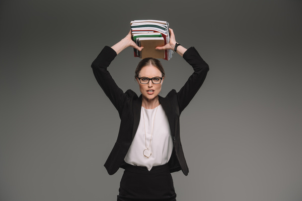 angry female teacher in eyeglasses holding stack of textbooks over head isolated on grey background  - Foto, Bild