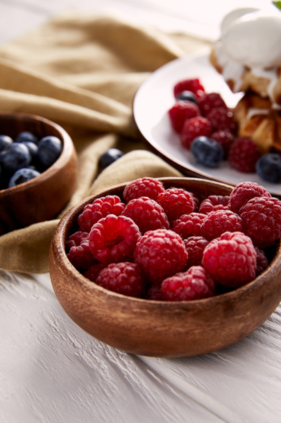 close-up shot of bowls with berries with belgian waffles blurred on background on white wooden table - Photo, Image