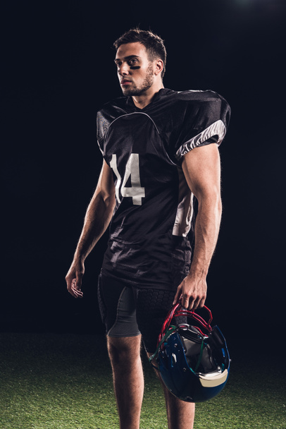 Αθλητικός αμερικανικός παίκτης ποδοσφαίρου στην μαύρη στολή κρατώντας το κράνος και να την βλέπουν μακριά μαύρη - Φωτογραφία, εικόνα
