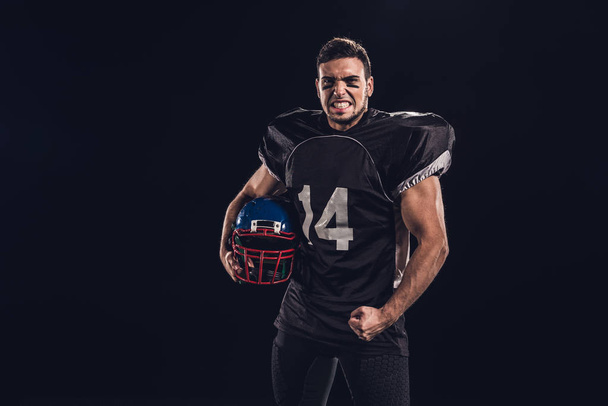 jugador de fútbol americano enojado en uniforme negro con casco y mirando a la cámara aislada en negro
 - Foto, Imagen