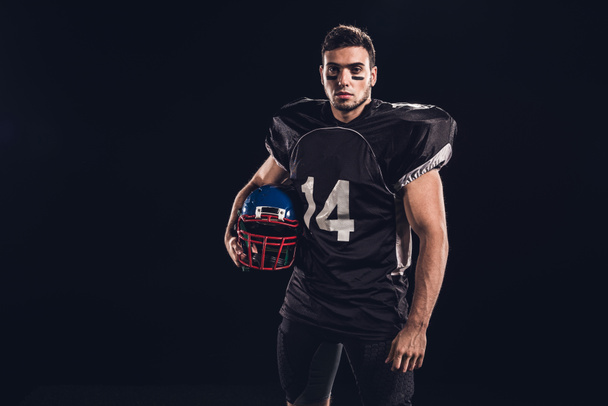 atlético jugador de fútbol americano en uniforme negro con casco y mirando a la cámara aislada en negro
 - Foto, imagen