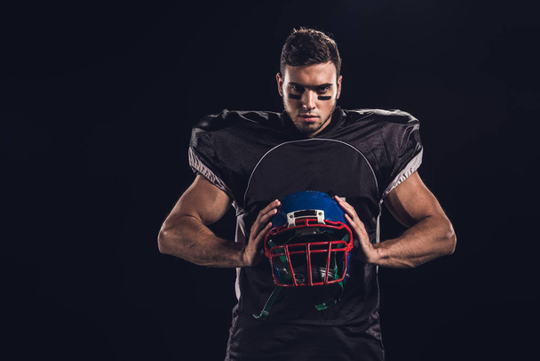 hráč amerického fotbalu v rukou držel helmu a při pohledu na fotoaparát izolované na černém - Fotografie, Obrázek