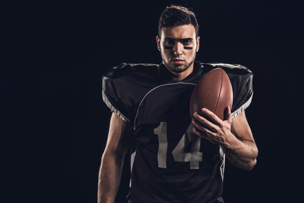 joven jugador de fútbol americano serio con bola mirando a la cámara aislada en negro
 - Foto, imagen