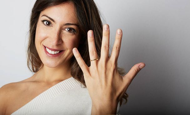 Happy european girl with long dark hair in stylish clothes smiling and holding hand on hip while showing trendy wedding ring over empty gray wall background.Copy paste text message space. Close-up. - Photo, image