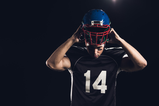 νεαρός ποδοσφαιριστής Αμερικανός τοποθέτηση στο κράνος που απομονώνονται σε μαύρο - Φωτογραφία, εικόνα