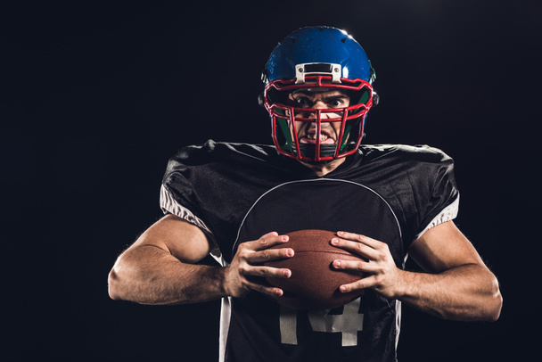 gekke american football speler bal in handen houden en kijken naar camera geïsoleerd op zwart - Foto, afbeelding